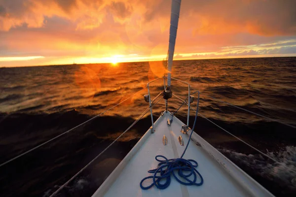 White yacht sailing in an open sea at sunset. A view from the deck to the bow, mast, sails. Epic cloudscape. Dramatic sky with glowing golden clouds after the storm. Racing, sport, leisure activity