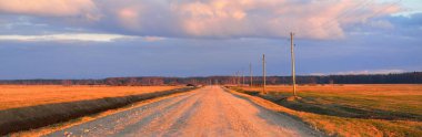 Gün batımında Letonya 'da tarladan geçen boş bir köy yolu. Transformatör direkleri yakın çekim. Arabadan bir manzara. Idyllic kırsal kesim. Renkli parlayan bulutlar. Ulaşım, yolculuk, özgürlük kavramları