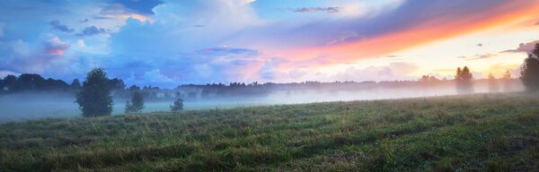 Epic colorful sunset sky and fog above the green plowed agricultural field, forest in the background. Dramatic cloudscape. Rural scene. Finland