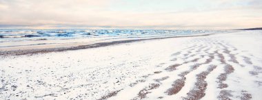 Gün batımında karla kaplı Baltık deniz kıyısından bir manzara. Riga Körfezi, Letonya. Renkli dramatik bulut manzarası. Kararsız hava, dalgalar ve su sıçramaları. Kış turizmi, küresel ısınma teması