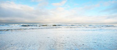 Gün batımında karla kaplı Baltık deniz kıyısından bir manzara. Riga Körfezi, Letonya. Dramatik bir gökyüzü. Kristal berrak mavi su. Kararsız hava, dalgalar ve su sıçramaları. Kış turizmi, küresel ısınma teması