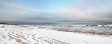 Gün batımında karla kaplı Baltık deniz kıyısından bir manzara. Riga Körfezi, Letonya. Renkli dramatik bulut manzarası. Kararsız hava, dalgalar ve su sıçramaları. Kış turizmi, küresel ısınma teması