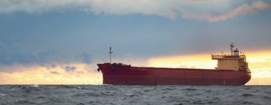 Büyük kırmızı kargo gemisi karanlık günbatımı göğünün altında açık denizde yol alıyor. Southampton 'da. İngiltere. Dramatik bulutlar, kasırga, fırtınalı hava. Küresel iletişim, nakliye, endüstri, lojistik