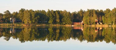 Kır evleri, yemyeşil ağaçlar ve Letonya 'da gün batımında bir nehir. Suyun simetri yansımaları. Sonbahar manzarası, huzurlu kırsal manzara. Çevre koruma, eko turizm teması