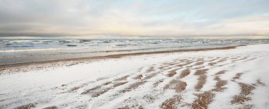 Gün batımında karla kaplı Baltık deniz kıyısından bir manzara. Riga Körfezi, Letonya. Renkli dramatik bulut manzarası. Kararsız hava, dalgalar ve su sıçramaları. Kış turizmi, küresel ısınma teması