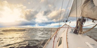 Gün batımında bir kış günü açık denizde yelken açmak. Güverteden pruvaya, direğe ve yelkenlere yakın görüntü. Dramatik gökyüzü, kara bulutların arasından geçen altın güneş ışığı. Destansı deniz manzarası. Kuzey denizi, Norveç