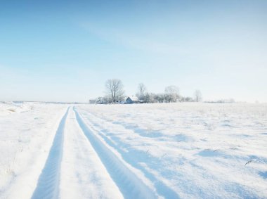 Güneşli bir günde kırsal alanda karla kaplı tek şeritli yol. Finlandiya. Açık mavi gökyüzü. Yaprak döken ve yemyeşil ağaçlar arka planda kırağı kaplamış. Idyllic kış manzarası
