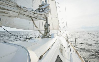 Güneşli bir yaz gününde açık denizde yelken açan beyaz bir yat. Güverteden pruva ve yelkenlere kadar yakın çekim. Dalgalar ve su sıçramaları. Sirus bulutları ve uçak izleri olan açık mavi bir gökyüzü. Hollanda