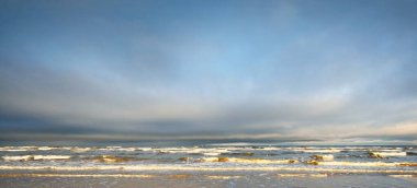 Gün batımında karla kaplı Baltık deniz kıyısından bir manzara. Riga Körfezi, Letonya. Dramatik mavi gökyüzü. Kararsız hava, dalgalar ve su sıçramaları. Kış turizmi, küresel ısınma teması