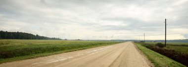 Yağmurdan sonra tarlalardan geçen boş bir köy yolu. Dramatik gökyüzü, destansı bulutlar. Kötü hava. Transformatör direkleri yakın çekim. Letonya