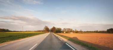 Gün batımında, Letonya 'da tarlalardan geçen boş bir otoyol. Arabadan bir manzara. Idyllic kırsal kesim. Ulaşım, yolculuk, özgürlük kavramları