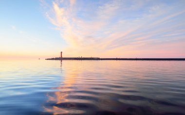 Gün batımında yağmurdan sonra Baltık Denizi. Pembe bulutları parlayan dramatik gökyüzü, suda simetri yansımaları. Arka planda deniz feneri var. Soyut doğal desen, doku, arkaplan, kavram sanatı