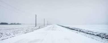 Kar fırtınasından sonra kırsal alanda karla kaplı bir yol. Elektrik hattı, trafo direkleri. Arabadan panoramik görüntü. Karanlık, fırtınalı bir gökyüzü. Off-road, lojistik, kış lastikleri, uzak köy