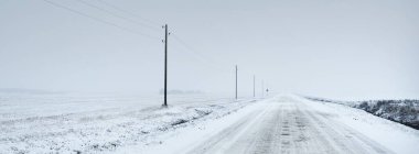 Kar fırtınasından sonra kırsal alanda karla kaplı bir yol. Elektrik hattı, trafo direkleri. Arabadan panoramik görüntü. Karanlık, fırtınalı bir gökyüzü. Off-road, lojistik, kış lastikleri, uzak köy