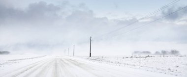 Tarlada karla kaplı kırsal yol. Elektrik hattı, trafo direkleri. Arabadan panoramik görüntü. Renkli bulutlar, dramatik günbatımı gökyüzü. Off-road, lojistik, kış lastikleri, uzak köy