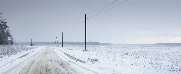 吹雪の後 フィールドを介して雪に覆われた田舎道 送電線 変圧器の棒 車からのパノラマビュー 暗い嵐の空 オフロード 冬のタイヤ リモート村 — ストック写真