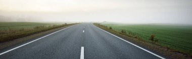 Gün doğumunda sisli bir havada boş otobanın tarlalar ve ormanlar arasındaki panoramik görüntüsü. Avrupa. Ulaşım, lojistik, seyahat, yolculuk, özgürlük, sürüş. Kırsal alan