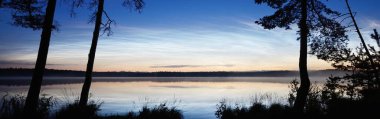 Yaz gündönümü gecesinde Saimaa Gölü 'nün üzerinde gece parlayan bulutlar ve sis vardı. Ağaç siluetleri. Altın güneş ışığı. Uzun pozlama. Destansı bulut manzarası. Suyun simetri yansımaları. Finlandiya