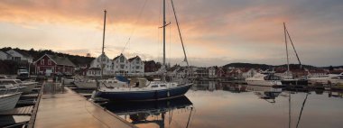 Mavi yelkenli gün batımında yat limanındaki bir rıhtıma demirlemiş. Renkli bulutlar, suya yansıyan yansımalar. Güney Norveç 'te bir köy. Tatil, spor, yelkencilik, yelkencilik, yelkencilik, yelkencilik, ulaşım, deniz aracı