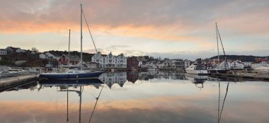 Mavi yelkenli gün batımında yat limanındaki bir rıhtıma demirlemiş. Renkli bulutlar, suya yansıyan yansımalar. Güney Norveç 'te bir köy. Tatil, spor, yelkencilik, yelkencilik, yelkencilik, yelkencilik, ulaşım, deniz aracı