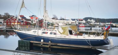 Mavi yelkenli gün batımında yat limanındaki bir rıhtıma demirlemiş. Renkli bulutlar, suya yansıyan yansımalar. Güney Norveç 'te bir köy. Tatil, spor, yelkencilik, yelkencilik, yelkencilik, yelkencilik, ulaşım, deniz aracı
