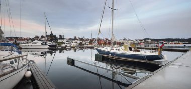 Mavi yelkenli gün batımında yat limanındaki bir rıhtıma demirlemiş. Renkli bulutlar, suya yansıyan yansımalar. Güney Norveç 'te bir köy. Tatil, spor, yelkencilik, yelkencilik, yelkencilik, yelkencilik, ulaşım, deniz aracı