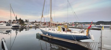 Mavi yelkenli gün batımında yat limanındaki bir rıhtıma demirlemiş. Renkli bulutlar, suya yansıyan yansımalar. Güney Norveç 'te bir köy. Tatil, spor, yelkencilik, yelkencilik, yelkencilik, yelkencilik, ulaşım, deniz aracı