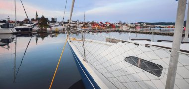 Mavi yelkenli gün batımında yat limanındaki bir rıhtıma demirlemiş. Renkli bulutlar, suda yansımalar. Yelkencilik, eğlence teması. Güney Norveç Köyü