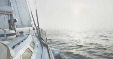 Güneş doğarken kalın bir siste, açık bir Baltık denizinde yelken açan beyaz bir yat. İsveç. Güverteden pruva ve yelkenlere kadar yakın çekim. Sabah güneşi ve parlayan bulutlar. Destansı deniz manzarası. Spor ve eğlence teması