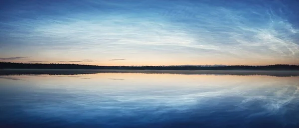 Starry sky with noctilucent clouds and fog above the Saimaa lake at summer solstice night. Tree silhouettes. Golden sunlight. Long exposure. Epic cloudscape. Symmetry reflections on the water. Finland