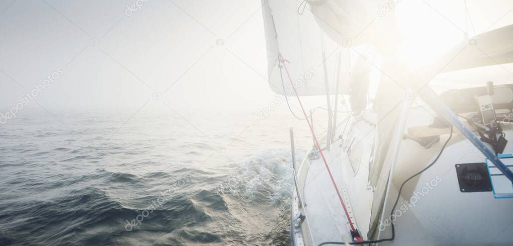 White yacht sailing in the Baltic sea. Thick fog. Sunrise. Sweden. Close-up view from the deck to the bow and sails. Morning sun, glowing clouds. Sport, recreation, leisure activity, cruise, regatta