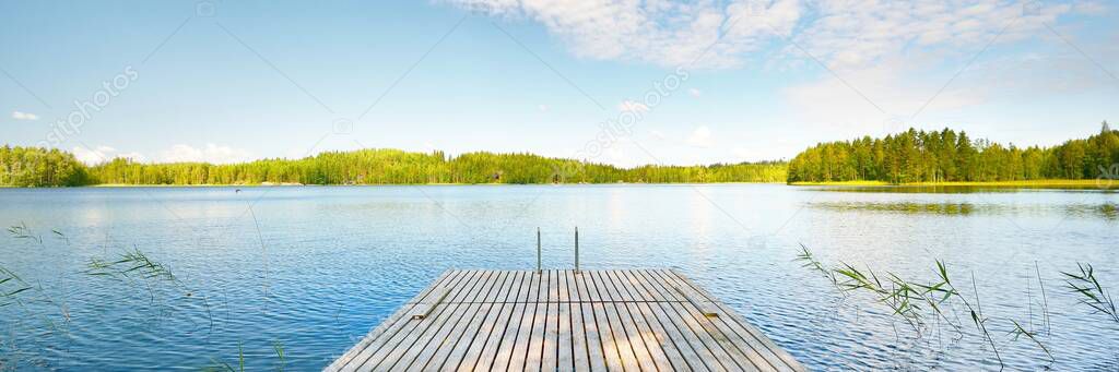 Wooden pier near the river close-up. Evergreen coniferous forest in the background. Clear blue sky, reflections on the water. Idyllic summer landscape. Eco tourism, recreation theme. Finland