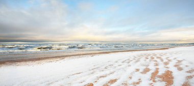Gün batımında karla kaplı Baltık deniz kıyısından bir manzara. Riga Körfezi, Letonya. Renkli dramatik bulut manzarası. Kararsız hava, dalgalar ve su sıçramaları. Kış turizmi, küresel ısınma teması