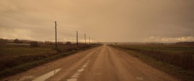 Gün batımında yağmurdan sonra tarlalardan geçen boş bir köy yolu. Dramatik renkli gökyüzü, destansı bulutlar. Transformatör direkleri yakın çekim. Letonya