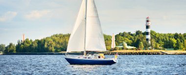 Gün batımında Riga körfezinde mavi yelkenli yelkenli yelkenli. Yakın plan. Baltık Denizi, Letonya. Arka planda deniz feneri var. Yarış, spor, eğlence, eğlence aktivitesi, takım çalışması kavramları.