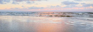 Gün batımında deniz kıyısında. Açık mavi gökyüzü, parlak pembe bulutlar, yumuşak ışık. Suyun simetri yansımaları, doğal ayna, su yüzeyi dokusu. Hava, iklim değişikliği, doğa. Idyllic deniz manzarası