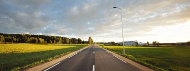 Gün batımında yeşil tarlalar ve huş ağacı ormanları boyunca boş bir otoyol (yeni asfalt yol). Sokak lambaları yakın çekim. Dramatik parlayan bulutlar. Seyahat yerleri, tatiller, sürüş, özgürlük
