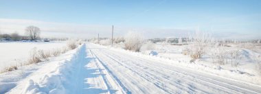 Güneşli bir günde kırsal alanda karla kaplı tek şeritli yol. Finlandiya. Açık mavi gökyüzü. Yaprak döken ve yemyeşil ağaçlar arka planda kırağı kaplamış. Idyllic kış manzarası