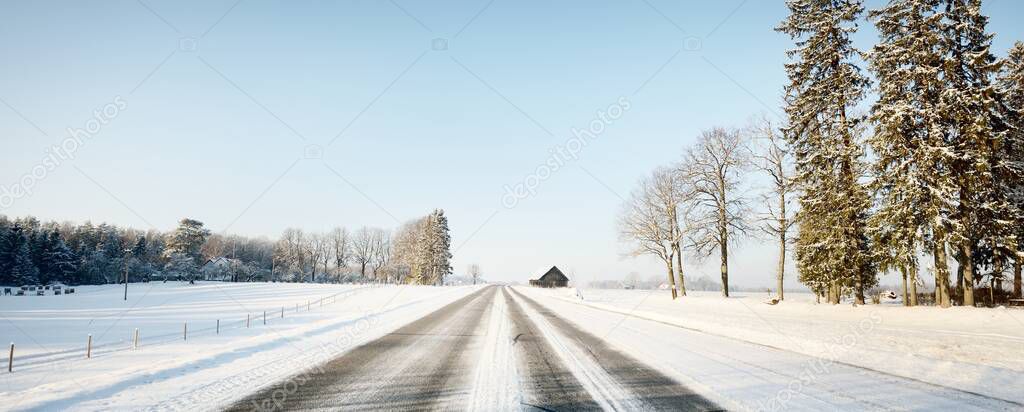 An empty asphalt road after cleaning. An old traditional house (log cabin) close-up. Car tracks in a fresh snow. Snow-covered forest and field in the background. Clear blue sky. Winter driving. Latvia