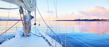 Gün batımında durgun suda yüzen beyaz bir yat. Buz ve güvertedeki ilk kar, pruvaya, direğe, halatlara ve yelkenlere yakın görüş. Renkli kış bulutlarıyla açık mavi gökyüzü. Norveç