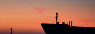 Büyük bir kargo gemisi gün batımında deniz fenerine doğru yola çıkıyor. Renkli bulutlu bir manzara. Riga Körfezi, Baltık Denizi, Letonya. Uluslararası iletişim, lojistik tema