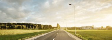 Gün batımında yeşil tarlalar ve huş ağacı ormanları boyunca boş bir otoyol (yeni asfalt yol). Sokak lambaları yakın çekim. Dramatik parlayan bulutlar. Seyahat yerleri, tatiller, sürüş, özgürlük