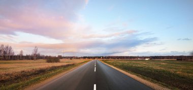 Boş bir otoyol (asfalt yol) yeşil kırsal tarım tarlaları gün batımında sürüldü. Güzel pembe bulutlar. Sıcak kış. İklim değişikliği teması. Letonya