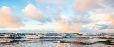 Gün batımında deniz kıyısında. Açık mavi gökyüzü, parlak pembe bulutlar, yumuşak ışık. Suyun simetri yansımaları, doğal ayna, su yüzeyi dokusu. Hava, iklim değişikliği, doğa. Idyllic deniz manzarası