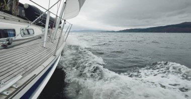 Bulutlu bir günde ahşap meşe güverteli, yelkenli, modern bir yat. Direğe ve yelkenlere bir manzara. Su, yakından sıçrıyor. Tarbert, İskoçya, İngiltere