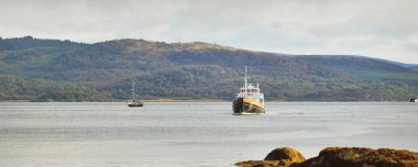 Bulutlu bir sonbahar günü. Sudan Tarbert kıyılarının manzarası. Arka planda orman var. Yat ve balıkçı teknesi yakın plan. İskoçya, İngiltere