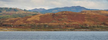 Sudan kayalık nehir kıyılarının panoramik görüntüsü. Arka planda ağaçlar, tepeler ve dağlar var. Bulutlu mavi gökyüzü. Gare Loch, Clyde Firth, İskoçya, İngiltere