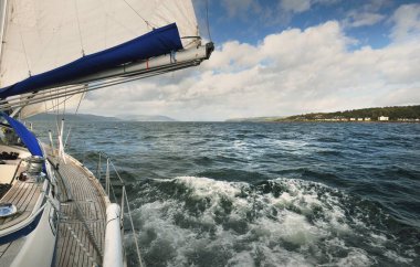 Bulutlu bir günde ahşap meşe güverteli, yelkenli, modern bir yat. Direğe ve yelkenlere bir manzara. Sudan kayalık nehir kıyılarının panoramik görüntüsü. Arka planda kır evleri, ağaçlar, tepeler ve dağlar var. Gare Gölü, Clyde Firth, S