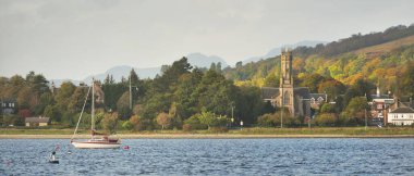 Güneşli bir günde yelkenli dolu bir yat. Arkasında tepeler, ormanlar ve kır evleri olan kayalık kıyılar. Rhu marinası, Clyde Firth, İskoçya, İngiltere