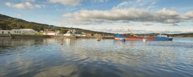 Küçük bir liman ve eski deniz fenerine boş bir gezinti güvertesi, kargo gemisine yakın çekim. Arka planda kayalık kıyılar var. Dramatik bulut manzarası. Ardrishaig, Crinan Kanalı, İskoçya, İngiltere